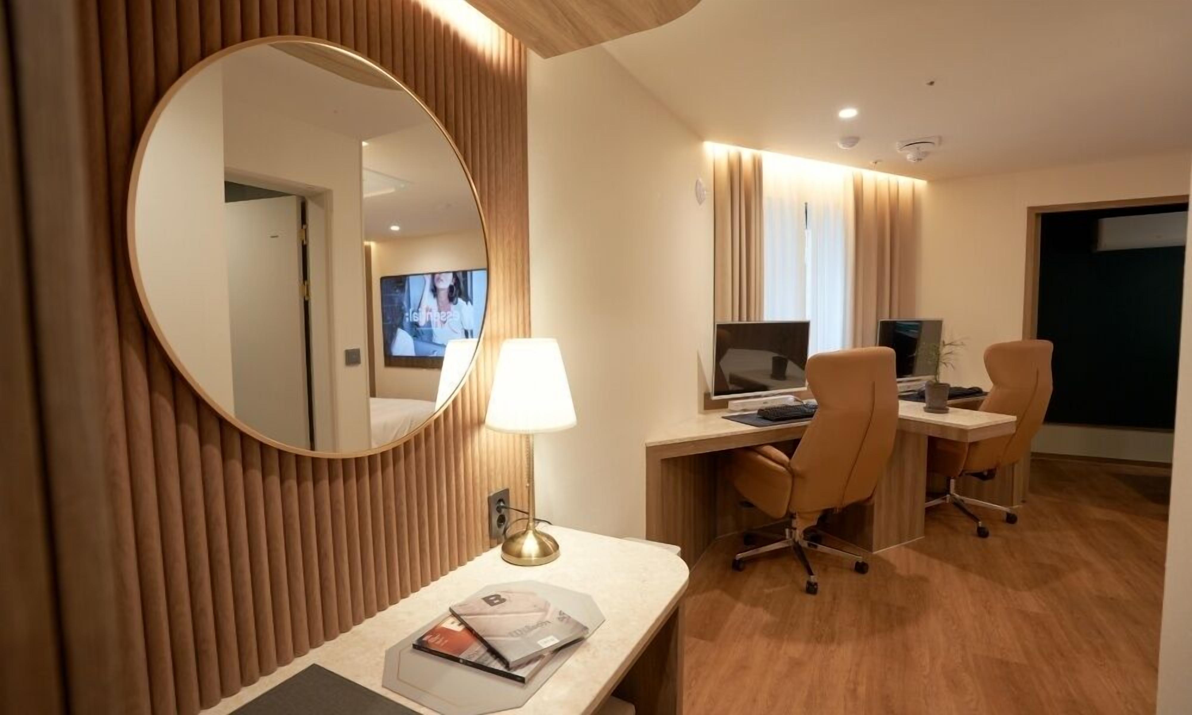 Une chambre d’hôtel avec un lambris en bois, un miroir rond, un bureau avec deux chaises, une télévision et une lampe.