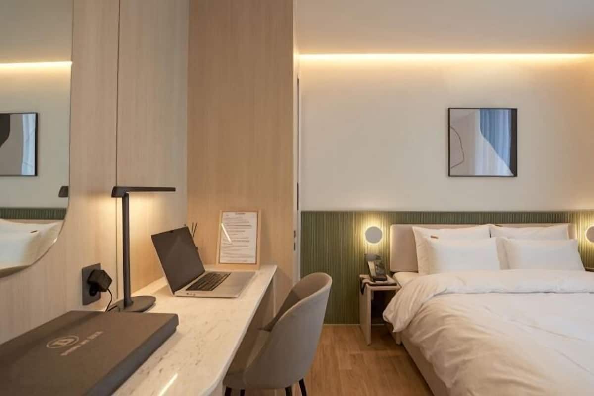 Een hotelkamer met een bed, een bureau met een laptop, een stoel, een nachtkastje met een lamp en een lijst aan de muur.