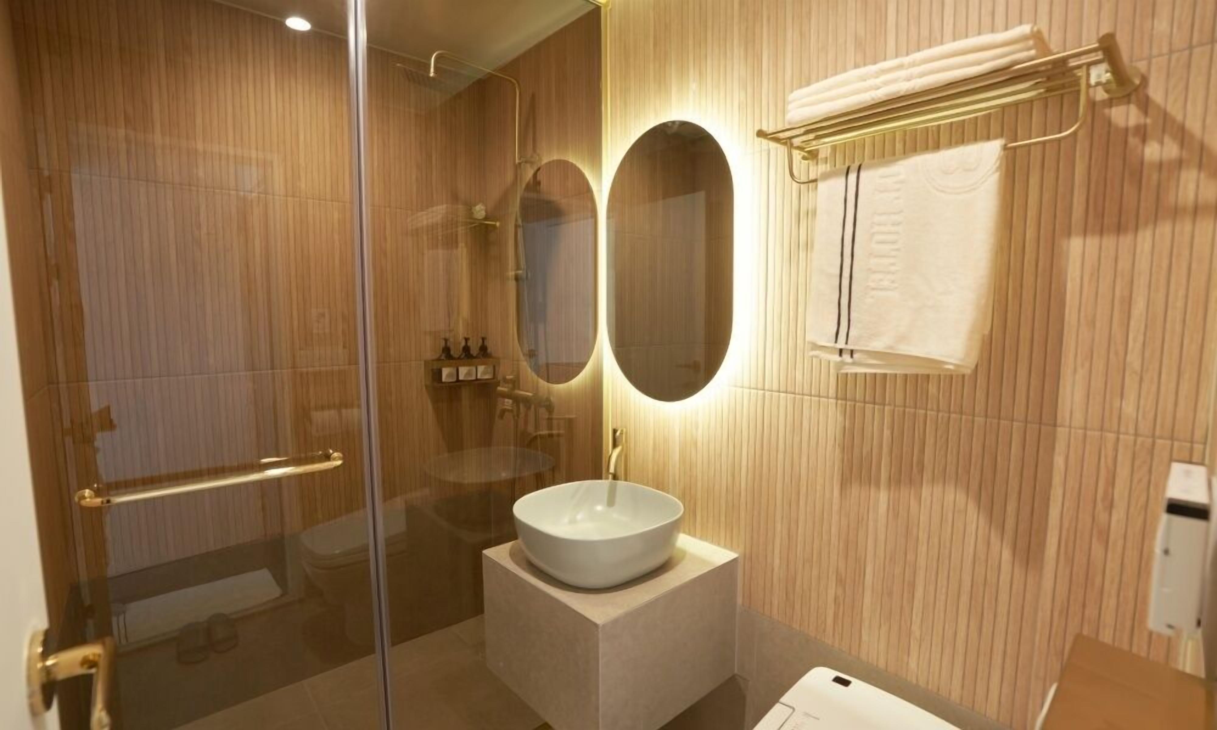 A modern bathroom with a glass shower enclosure, a round mirror, a white sink, and a towel rack.