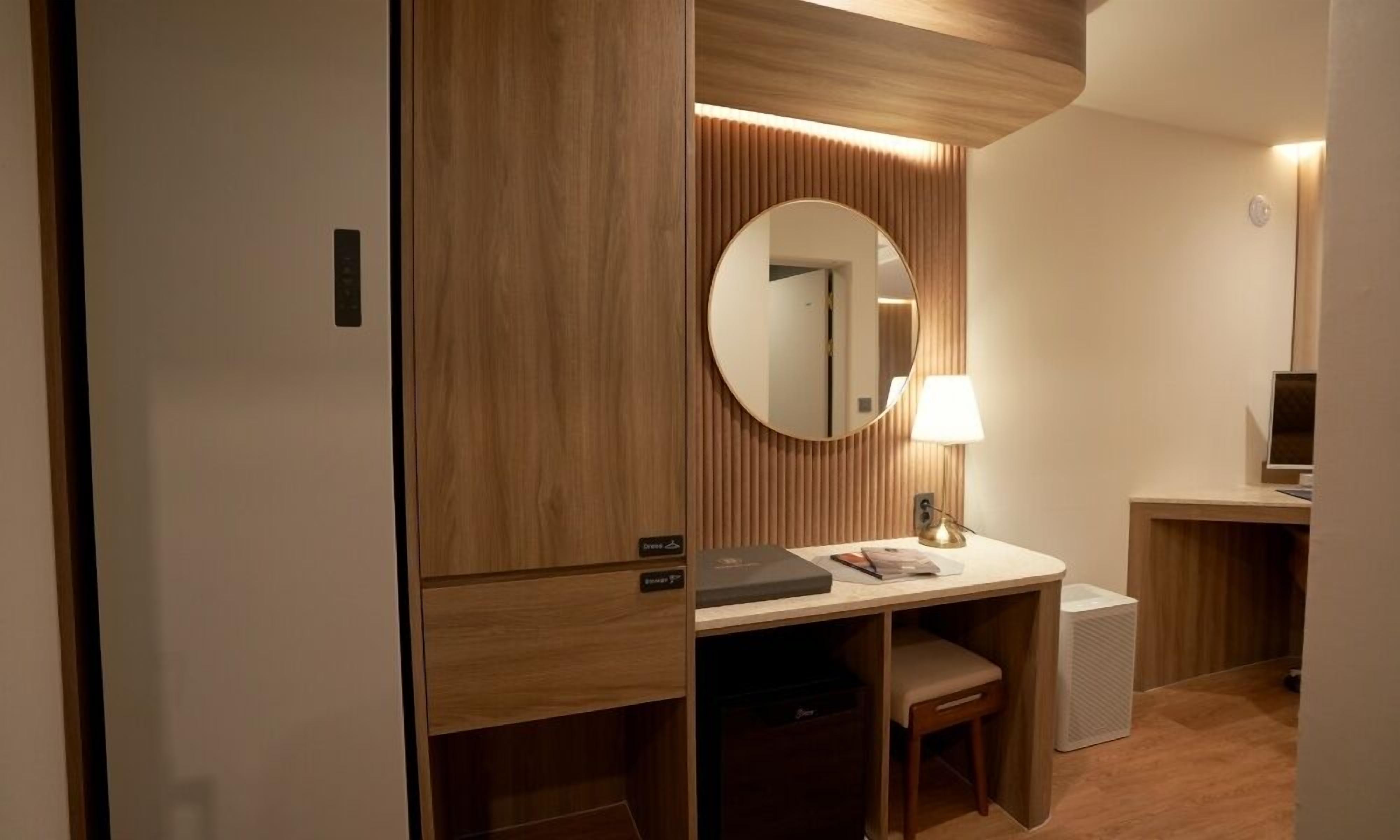 A hotel room with a wooden wardrobe, a desk with a lamp, a chair, and a round mirror.