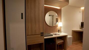 A hotel room with a wooden wardrobe, a desk with a lamp, a chair, and a round mirror.