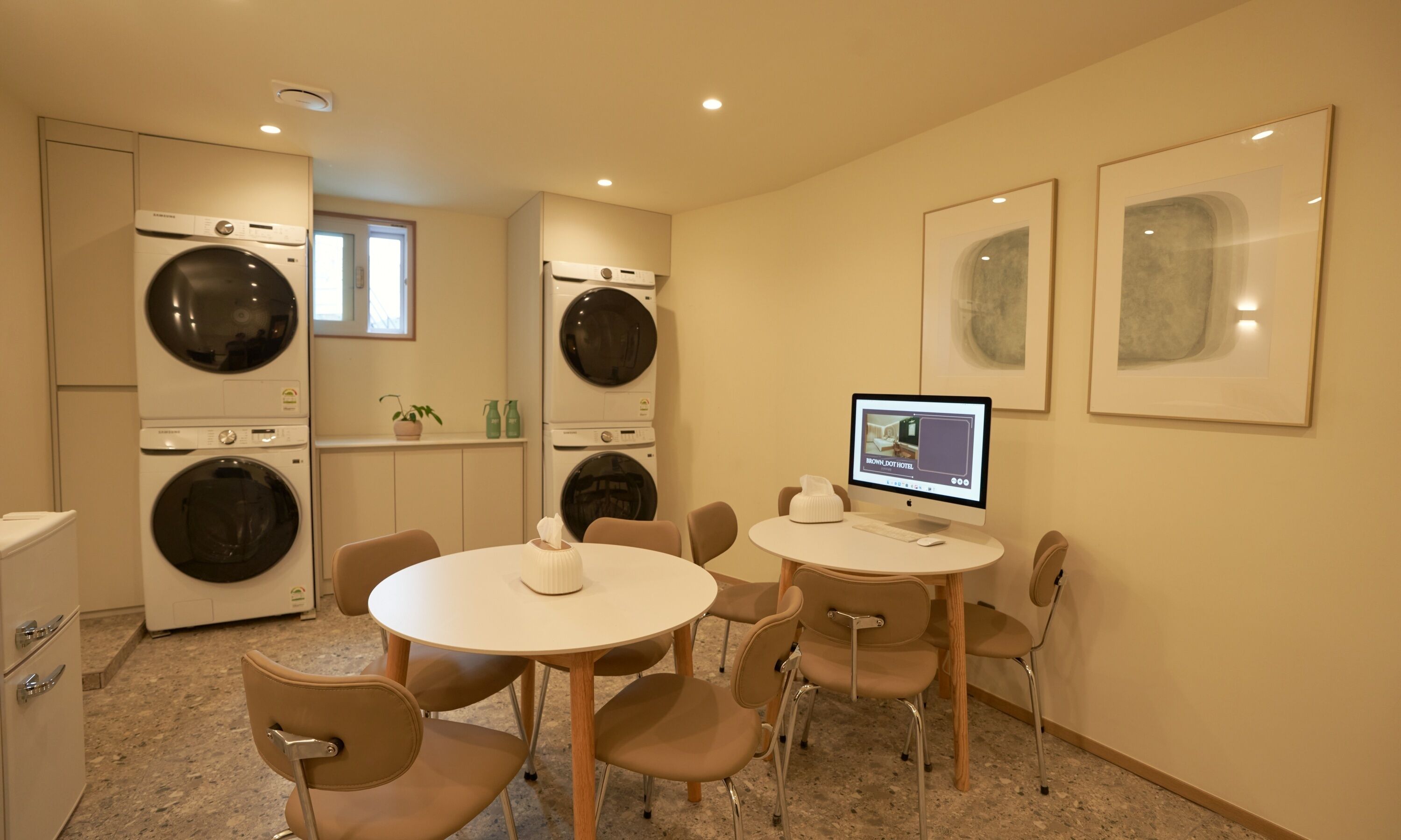 A laundry room with four washing machines, a table with chairs, and a computer monitor displaying a hotel room.