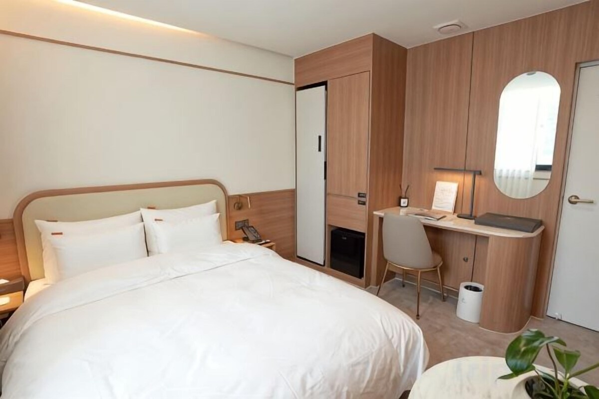 A hotel room with a bed, a desk, a chair, a mirror, and a trash bin.