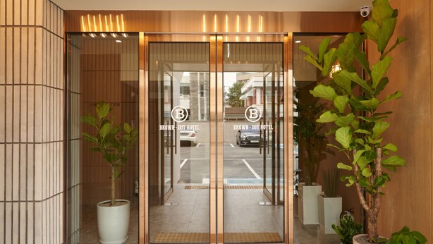 Entrada de hotel con diseño moderno, con puertas de vidrio que exhiben un logotipo, plantas en macetas y una cámara de seguridad.