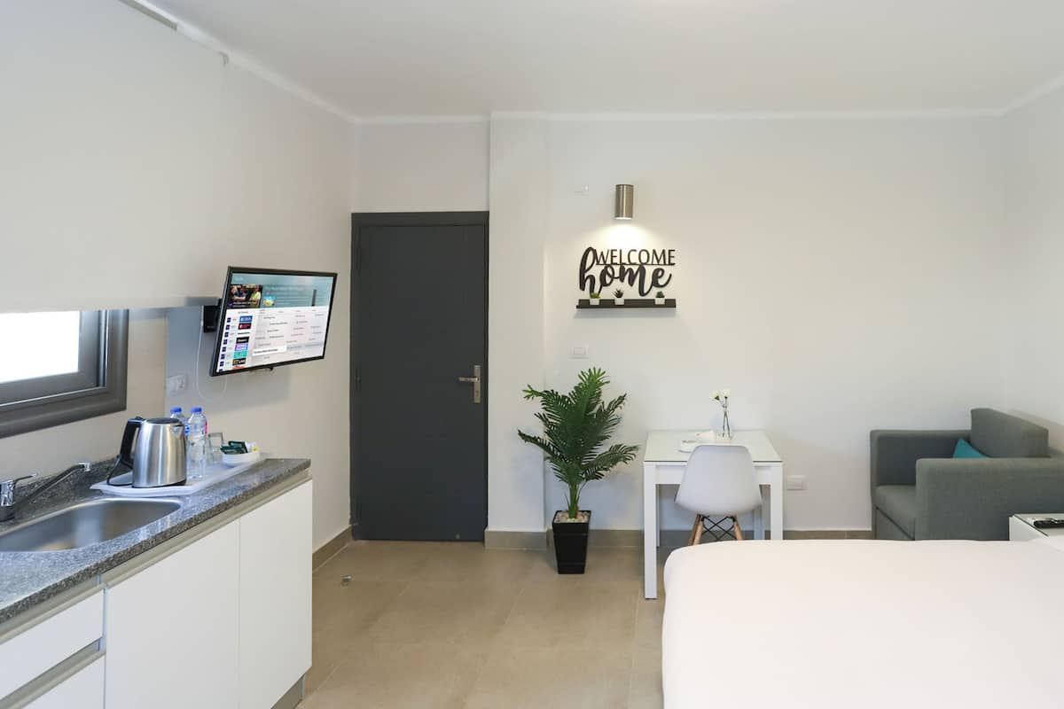 A modern hotel room with a bed, a desk, a sofa, a coffee machine, a TV, and a welcome sign.