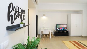 Un salon moderne avec une pancarte indiquant « BIENVENUE CHEZ VOUS », un téléviseur à écran plat, un bureau avec une chaise et une plante en pot.