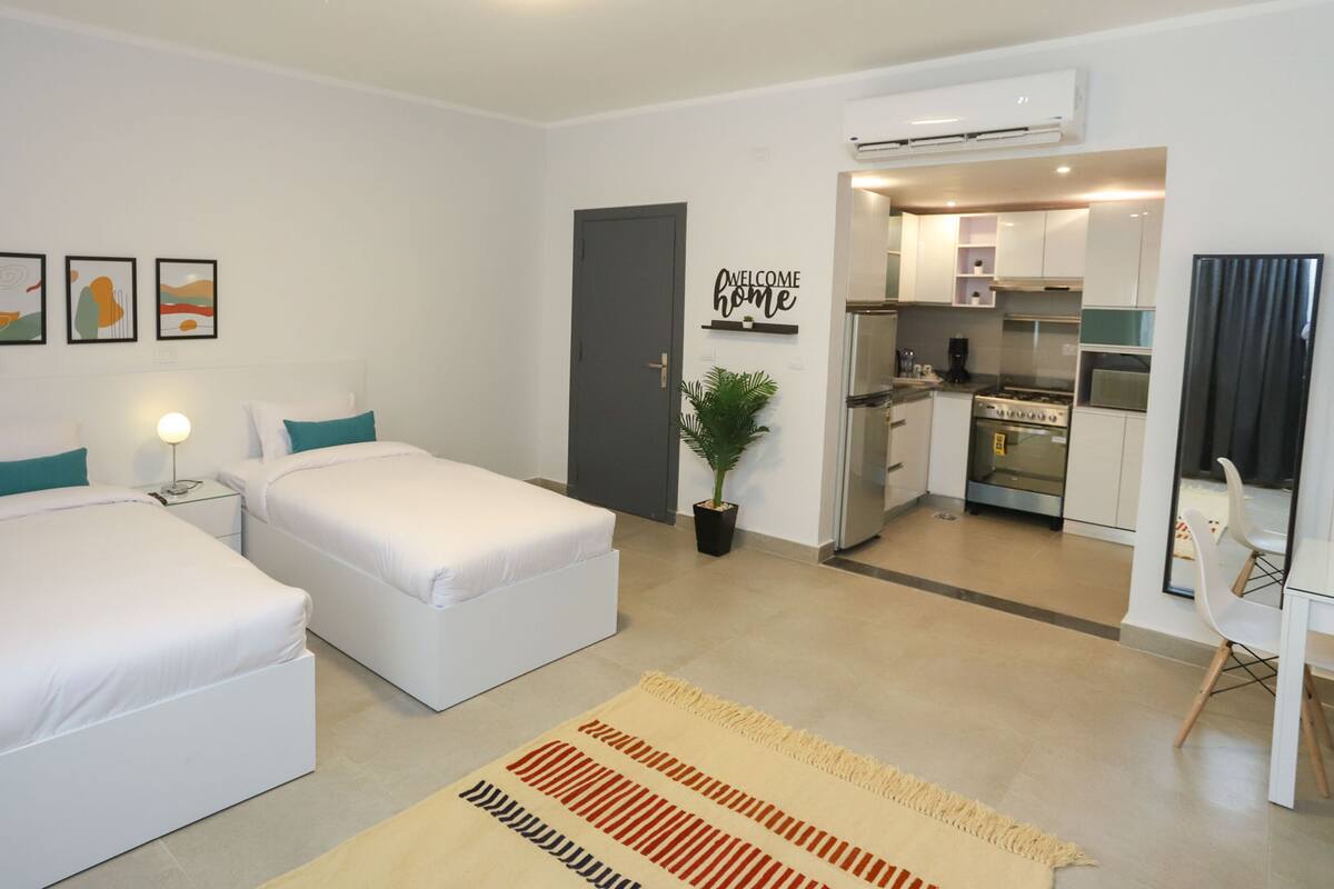 A modern hotel room with two single beds, a small bedside table with a lamp, a potted plant, and a kitchen area visible through an open door.