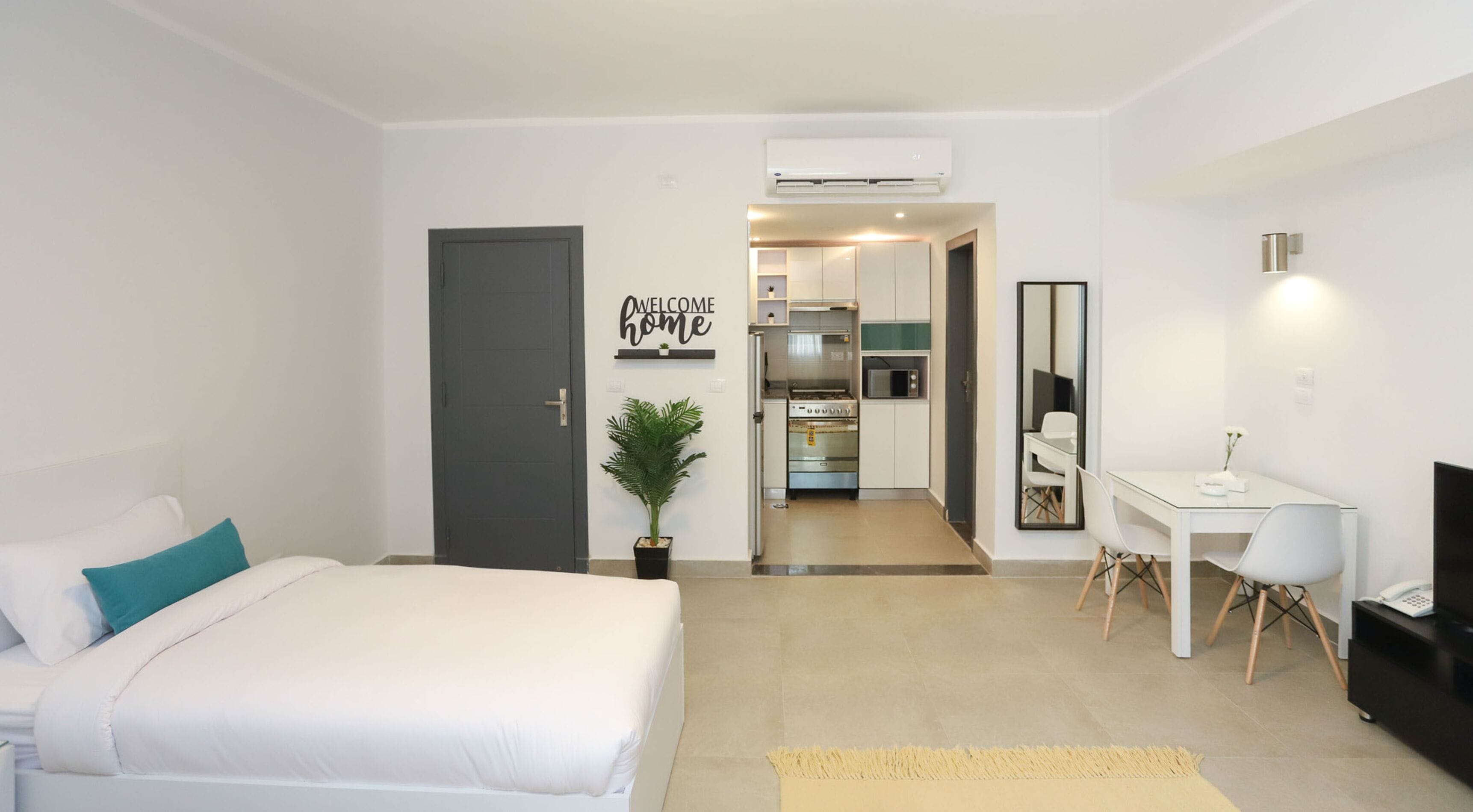 Une chambre d’hôtel moderne, avec un lit, un bureau, une chaise, une petite plante et un coin cuisine à l’arrière-plan.