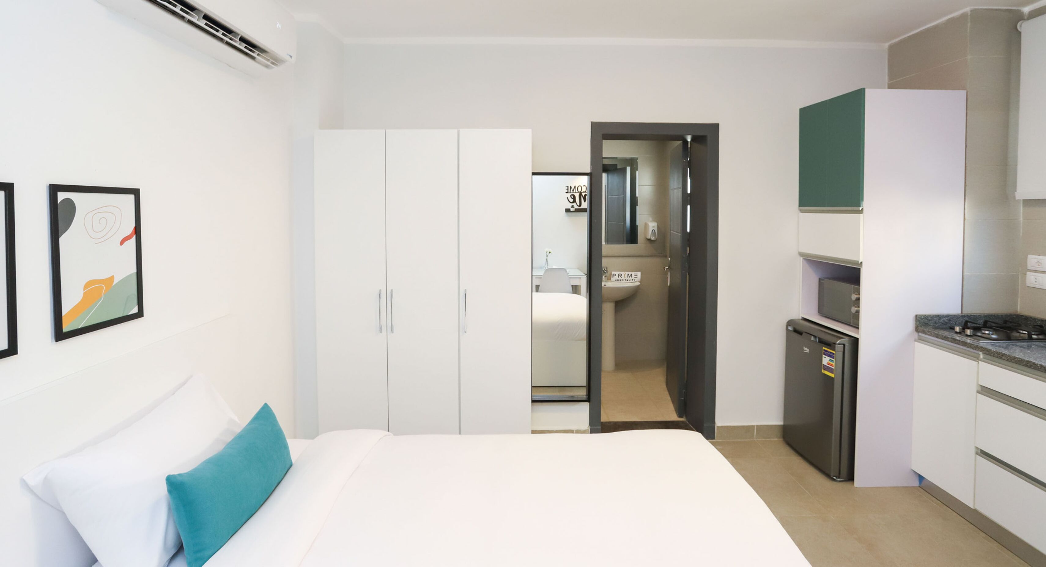 A hotel room with a bed, a wardrobe, a kitchenette, and a bathroom visible through an open door.