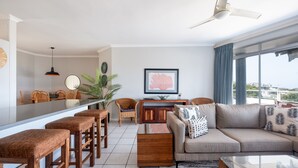 A modern living room with a sofa, coffee table, and bar stools. There is a dining area with chairs and a table, and a large window with curtains. - Barrington 116 (Ballito)
