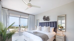 A spacious bedroom with a large bed, a ceiling fan, a nightstand, a mirror, and a view of the ocean. - Barrington 116 (Ballito)
