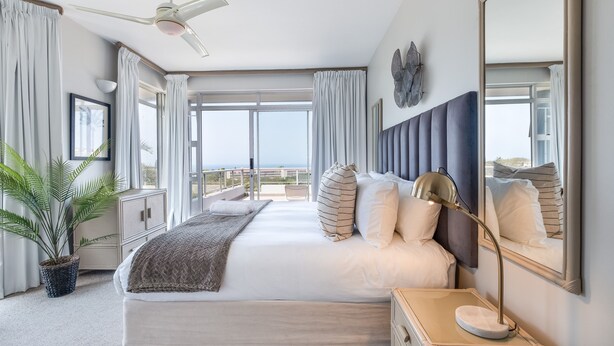 A spacious bedroom with a large bed, a ceiling fan, a bedside table with a lamp, and a view of the outdoors. - Barrington 116 (Ballito)
