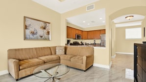 A living room with a sectional sofa, a glass coffee table, and a kitchen in the background.