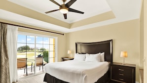 A hotel room with a large bed, a ceiling fan, a nightstand with a lamp, and a view of a golf course through a sliding glass door.