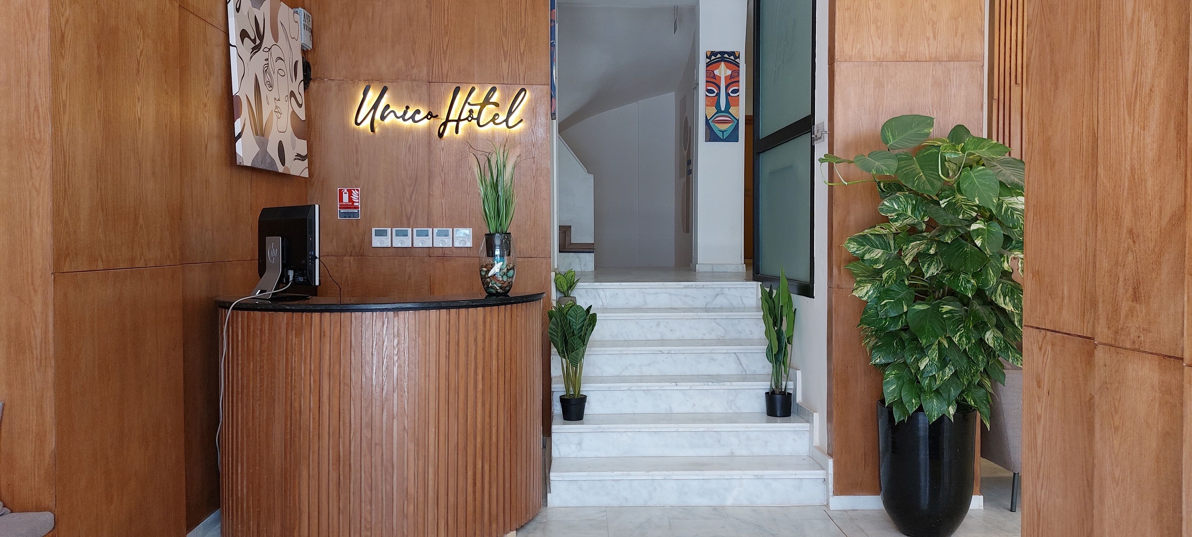 A hotel reception area with a wooden reception desk, a neon sign reading "Unico Hotel," and a staircase leading to the upper floor.