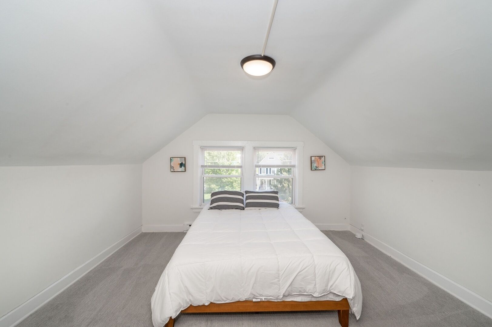 A bedroom with a sloped ceiling, a bed with white bedding, two windows with blinds, and a ceiling light fixture.