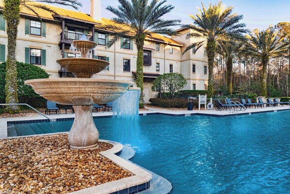 A swimming pool with a tiered fountain, surrounded by palm trees and a building with balconies.