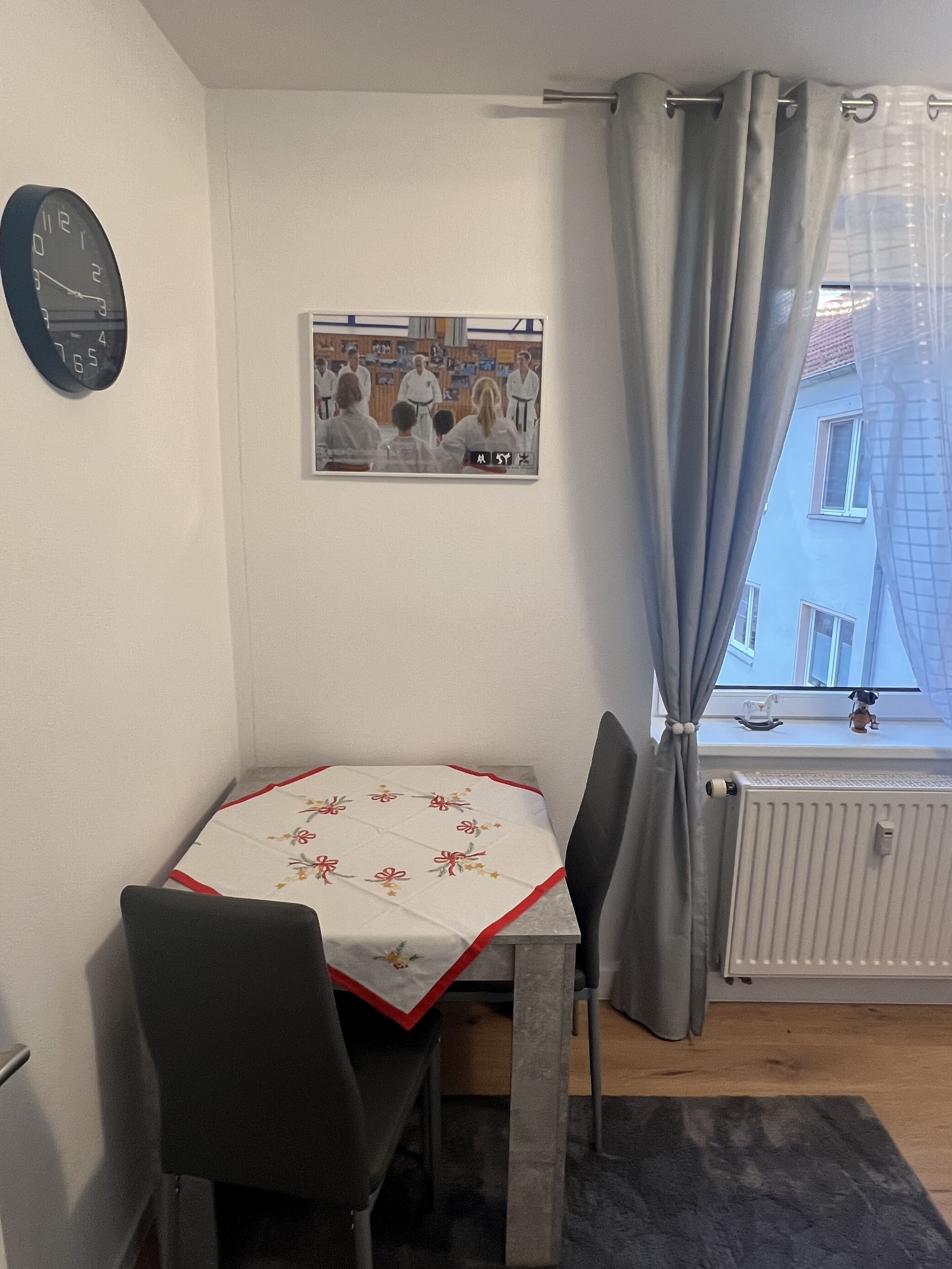Un petit coin repas avec une table recouverte d’une nappe à motifs floraux, deux chaises, une horloge au mur et une fenêtre avec des rideaux.