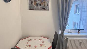 Un petit coin repas avec une table recouverte d’une nappe à motifs floraux, deux chaises, une horloge au mur et une fenêtre avec des rideaux.
