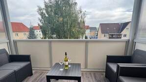 Ein Balkon mit Korbmöbeln, einem Tisch mit Flasche und Gläsern sowie Blick auf Gebäude und Bäume.