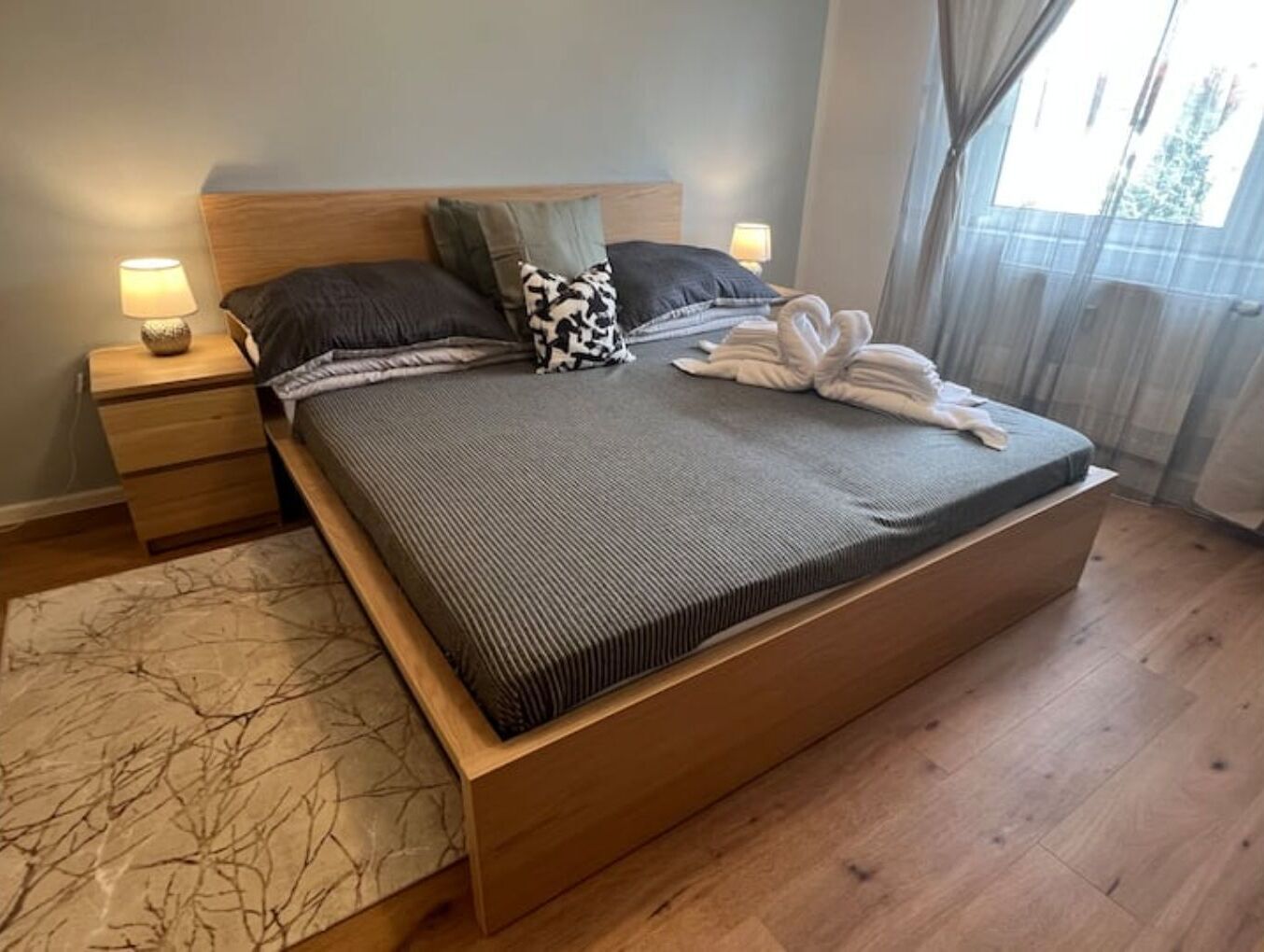 Ein großes Bett mit Holzrahmen, einer grauen Bettdecke und zwei Kissen. Ein Nachttisch mit Lampe und einer kleinen Dekoration steht daneben. Auf dem Bett liegt ein gefaltetes Handtuch.