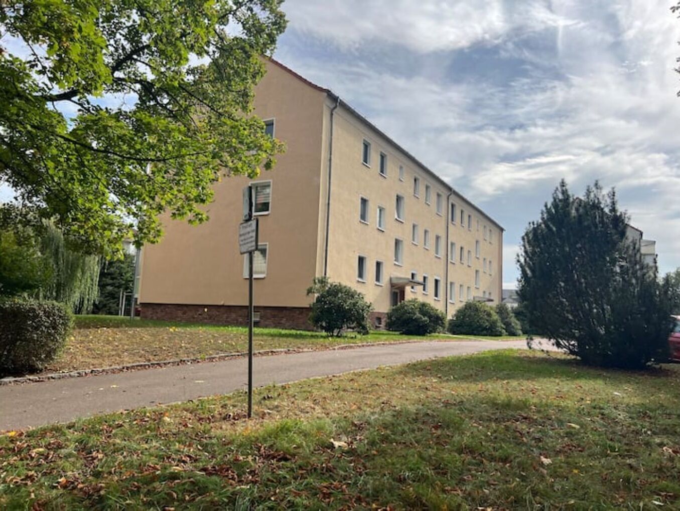 Un edificio de varios pisos con fachada color beige, un sendero pavimentado y una zona verde con árboles.