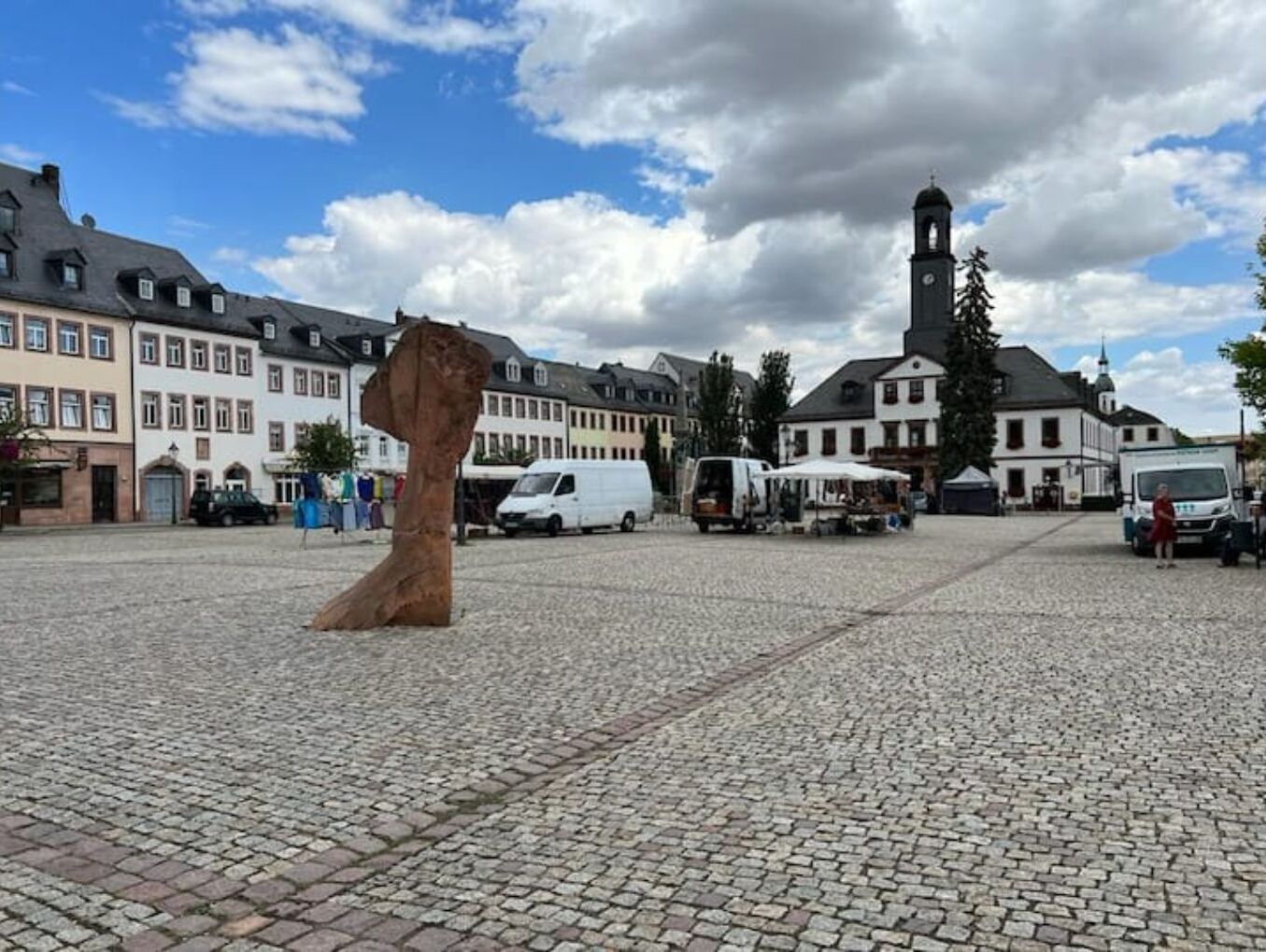 A cobblestone square with a large sculpture in the center, surrounded by historic buildings and a church tower in the background.