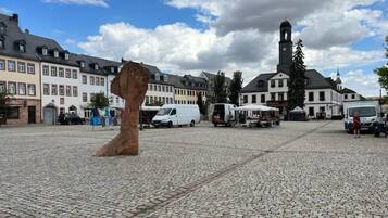 A cobblestone square with a large sculpture in the center, surrounded by historic buildings and a church tower in the background.