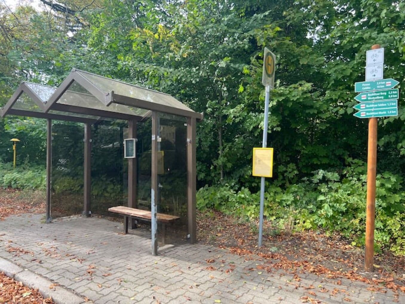 Ein überdachter Sitzplatz mit Bank, umgeben von Bäumen und einem gepflasterten Weg.