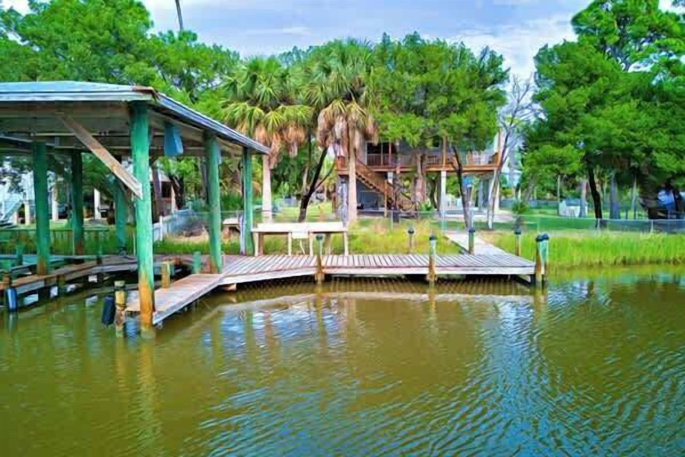 A wooden dock extending into a calm body of water, surrounded by lush greenery and a small pavilion.