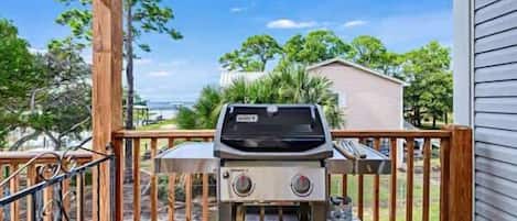 A barbecue grill on a wooden deck with a metal railing, a water container, and a house in the background.