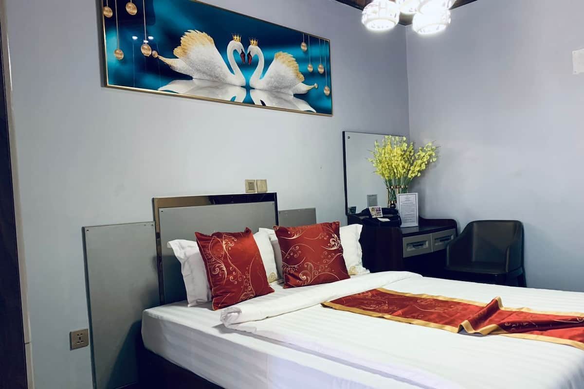 A hotel room with a large bed, a headboard, two red pillows, a white bedspread, a red throw blanket, a wall-mounted mirror, a desk with a chair, and a painting of swans.