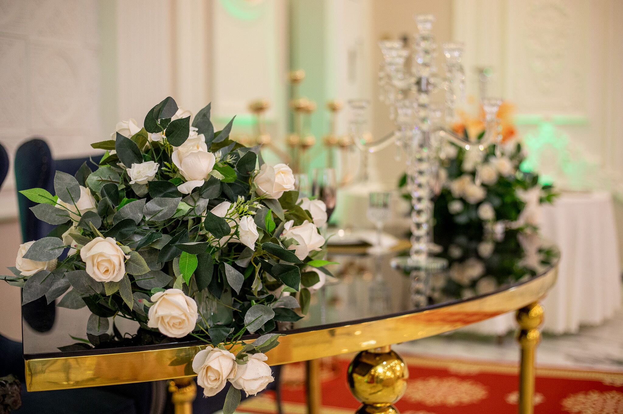 Una mesa con superficie de vidrio y patas doradas, decorada con rosas blancas y hojas verdes.