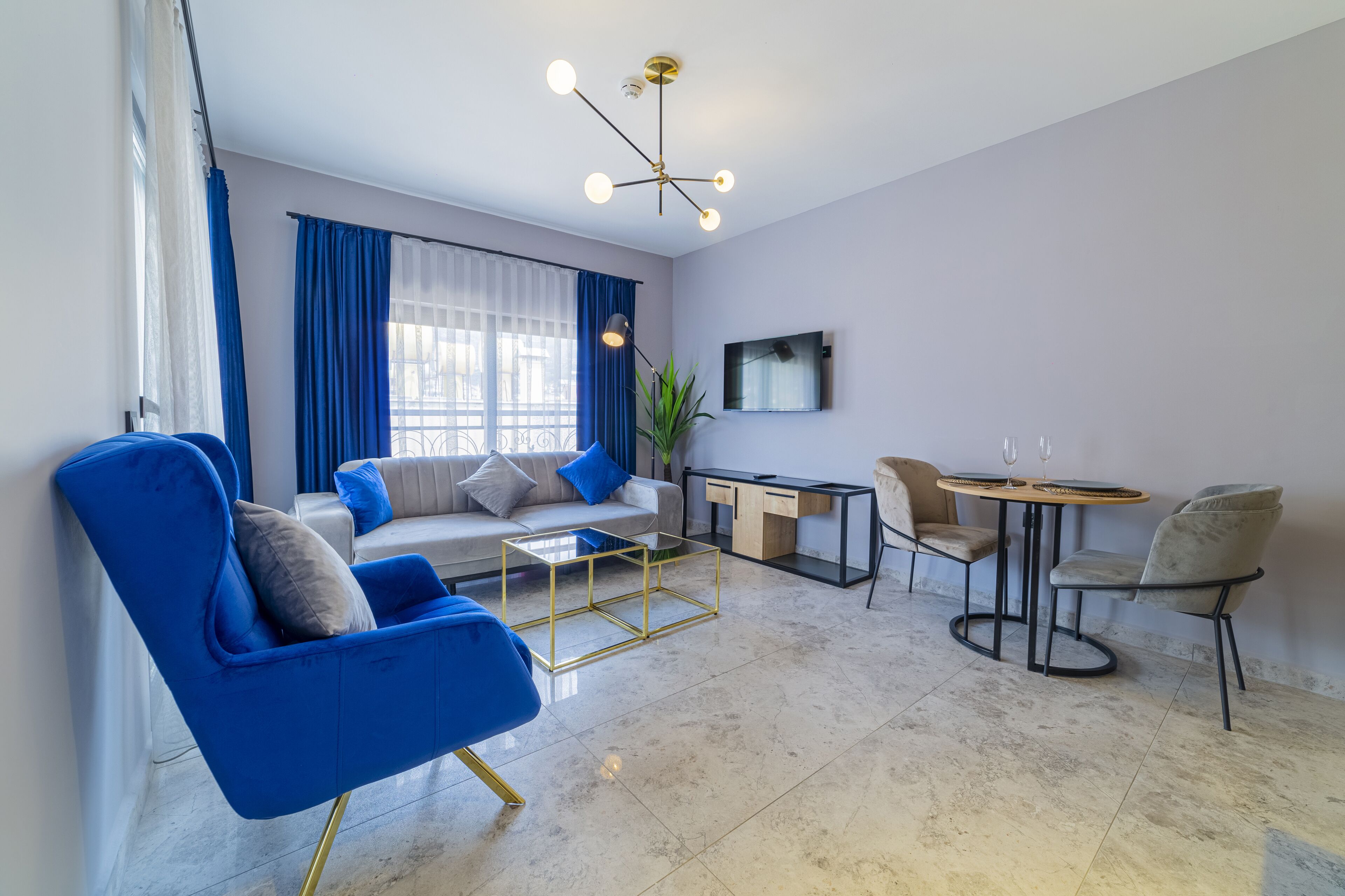 A modern living room with a blue armchair, a sofa, a coffee table, a dining table with chairs, a TV mounted on the wall, and a large window with blue curtains.