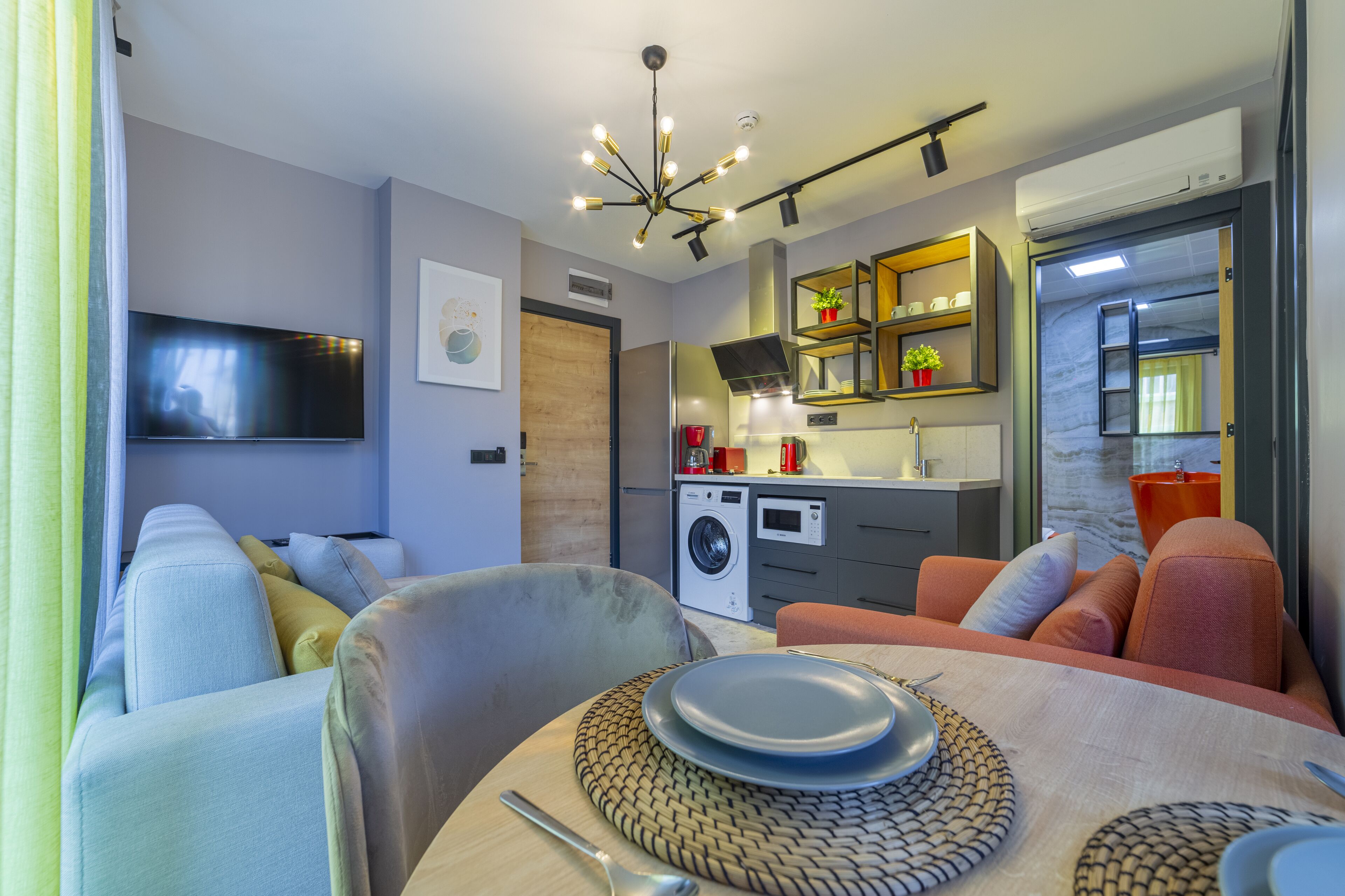 A compact living space with a sofa, dining table, kitchen area, and a TV mounted on the wall.