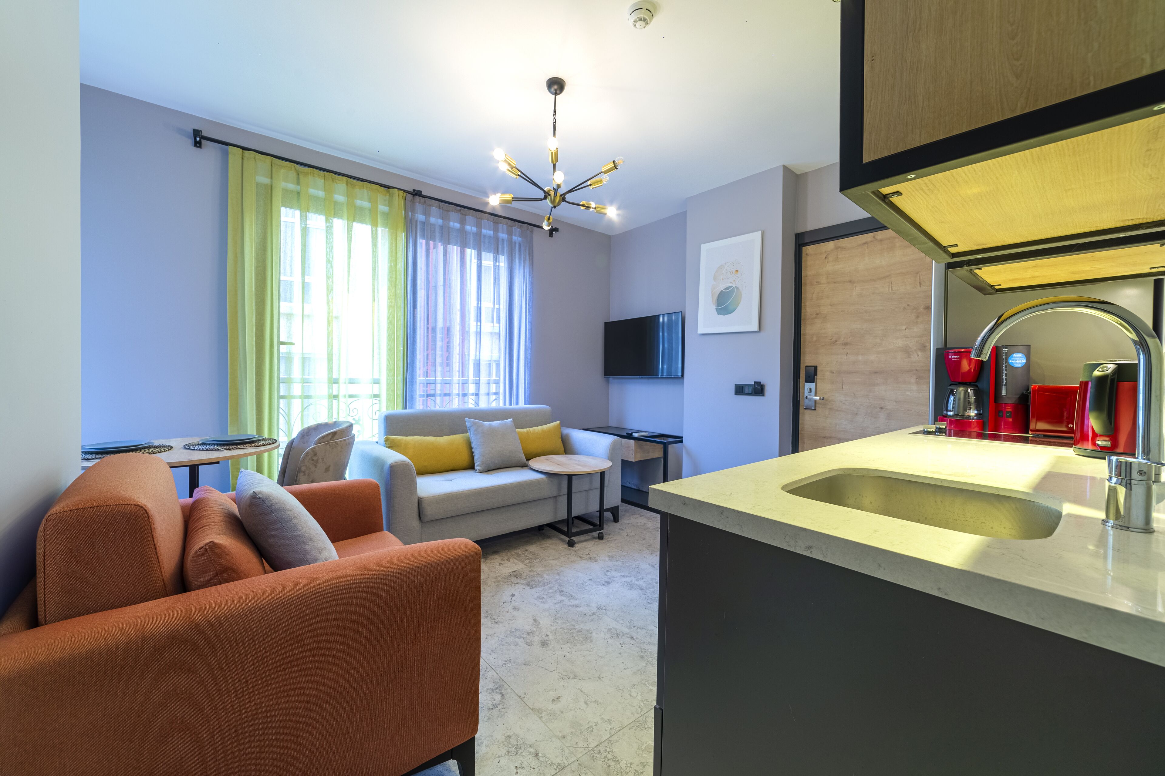 A modern living room with a sofa, a small round table, a TV mounted on the wall, and a kitchen area with a sink and red appliances.
