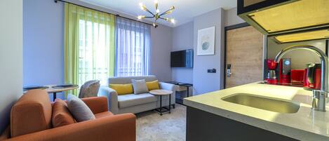 A modern living room with a sofa, a small round table, a TV mounted on the wall, and a kitchen area with a sink and red appliances.