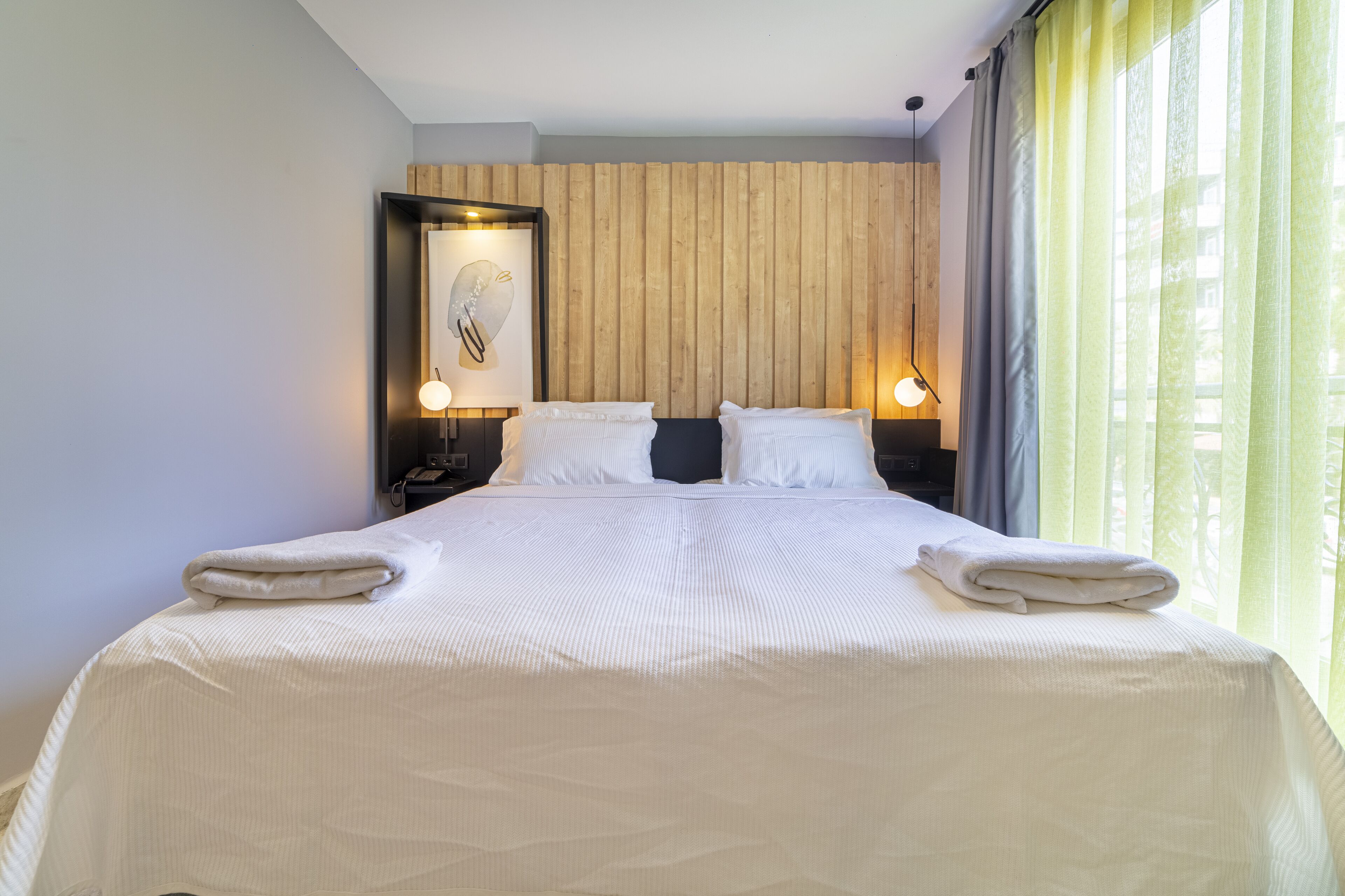 Una cama bien tendida con sábanas y toallas blancas, un cabecero de listones de madera y dos lámparas de noche.
