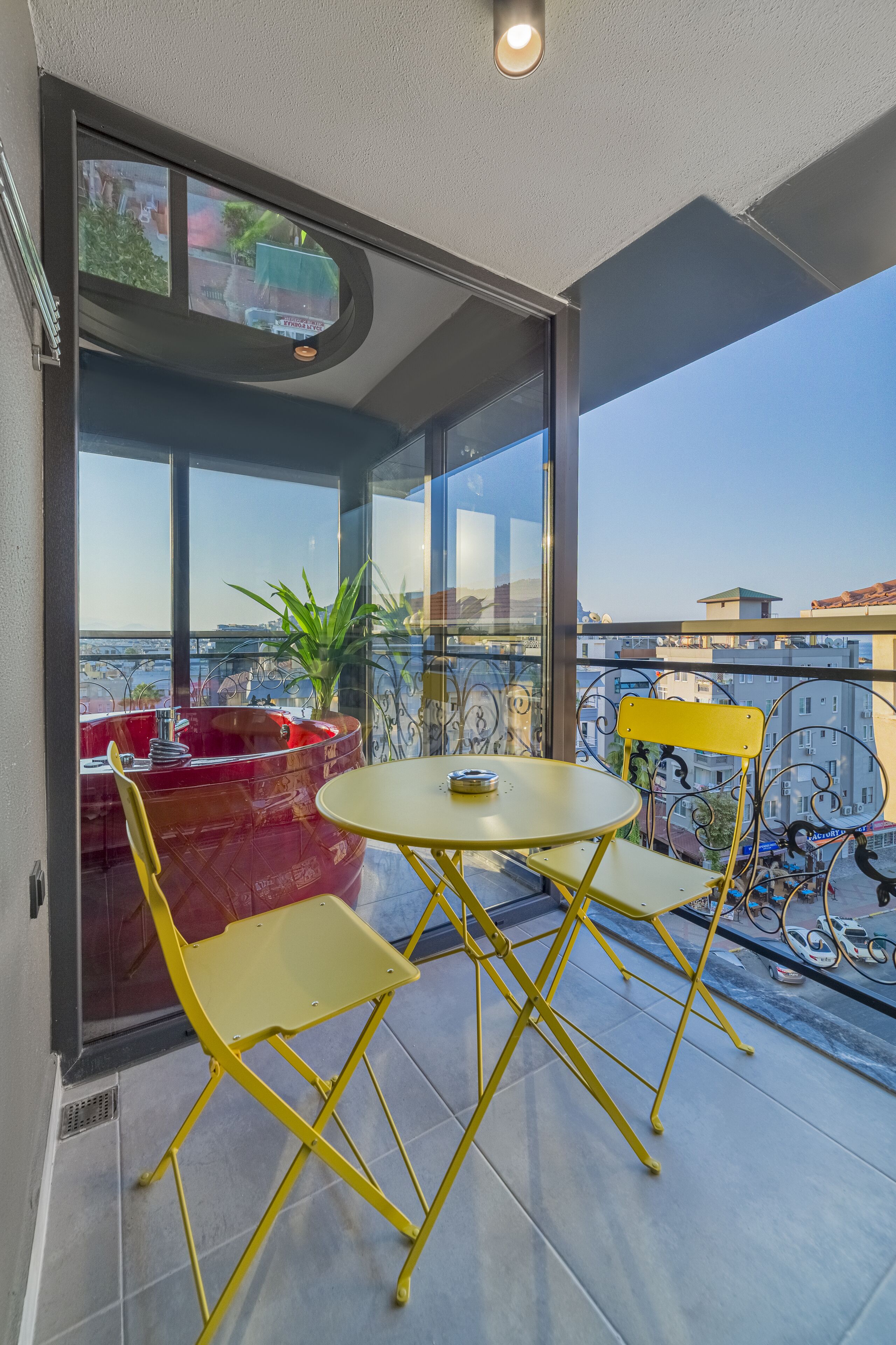 Un balcón con sillas plegables amarillas y una pequeña mesa redonda, ventanas amplias que ofrecen vistas a la ciudad y una planta en maceta.