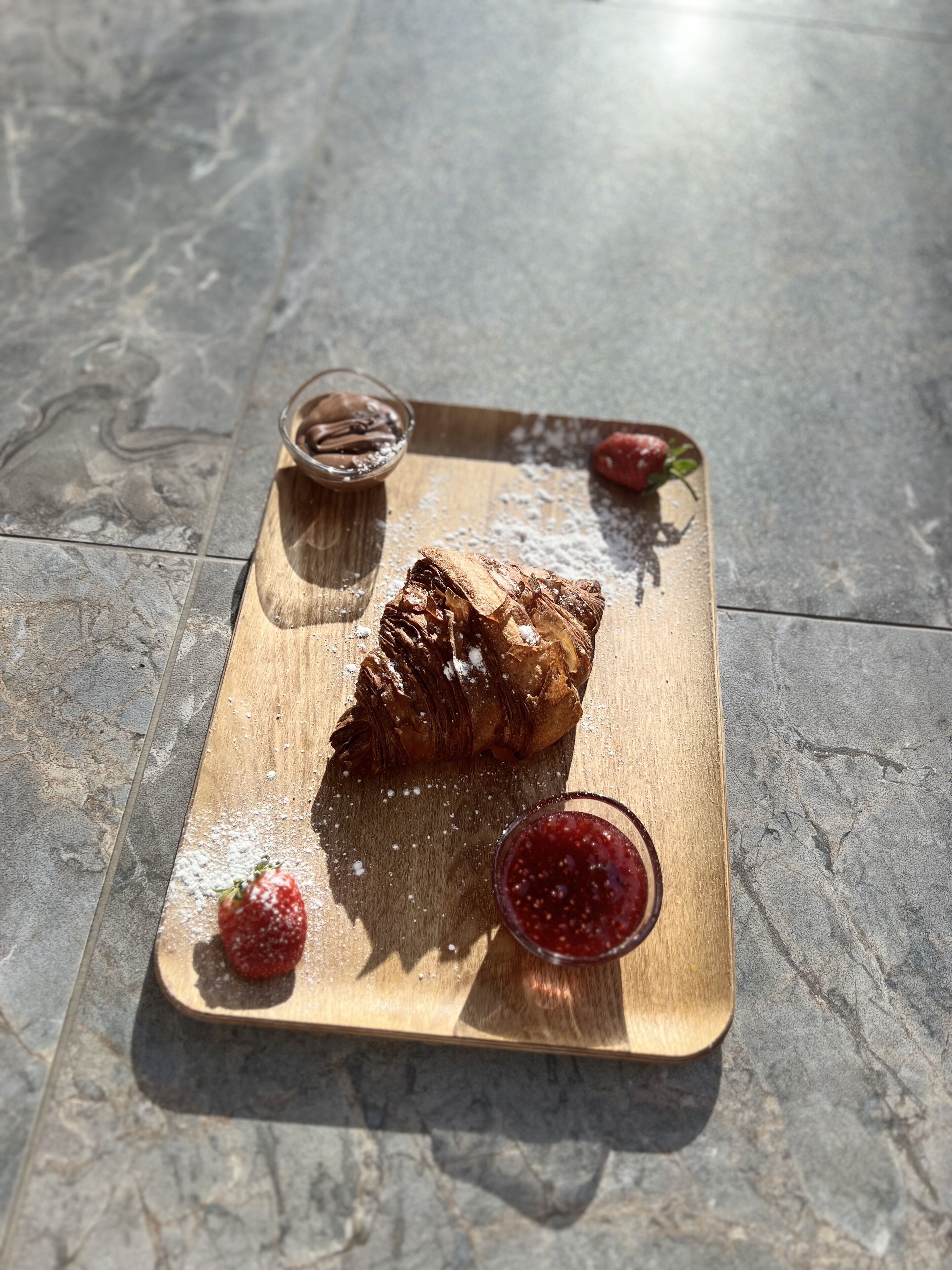 A croissant on a wooden board with powdered sugar, a strawberry, and a small bowl of jam.