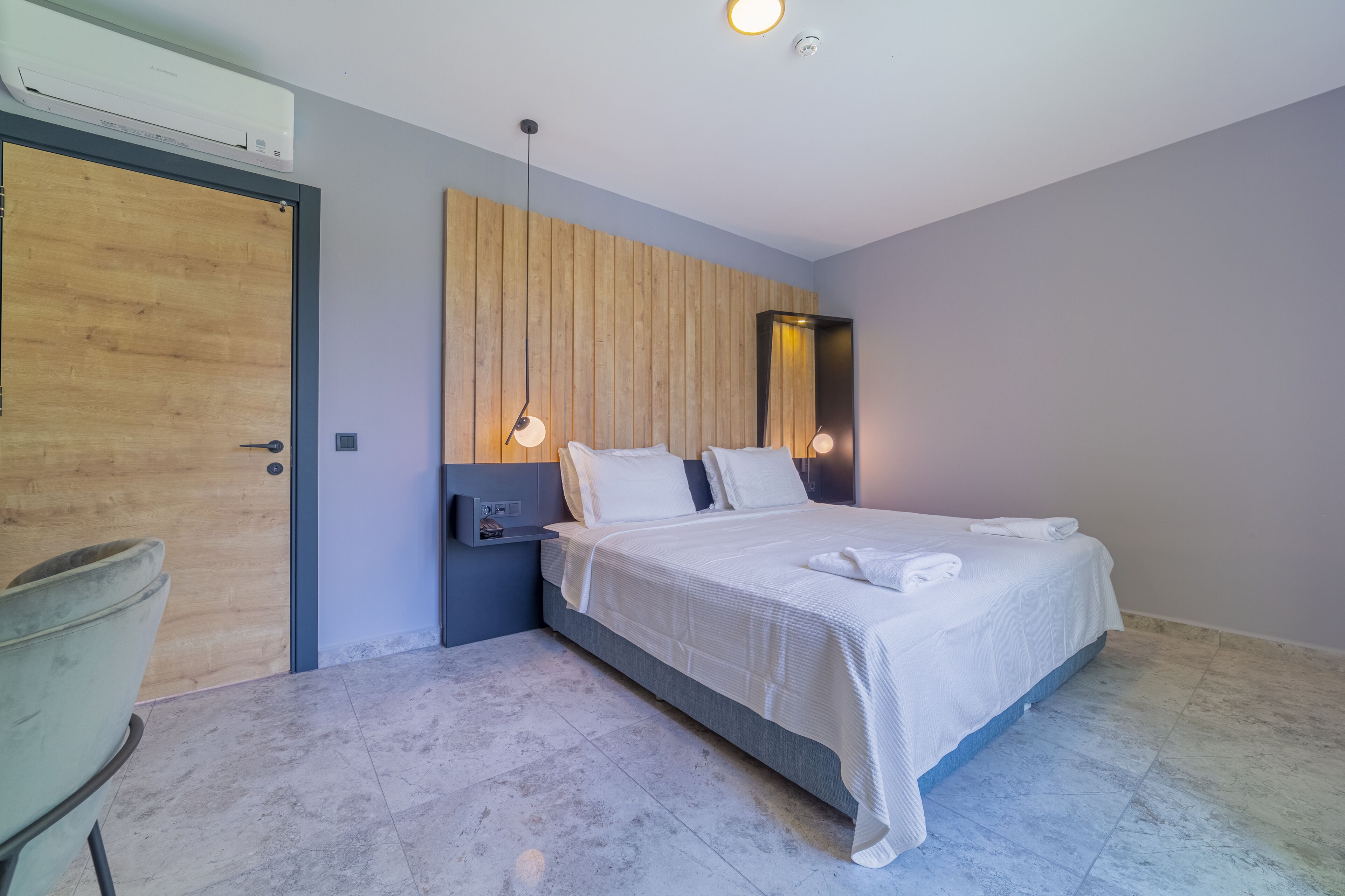 A modern hotel room with a wooden headboard, a bed with white linens, a nightstand, and a chair.