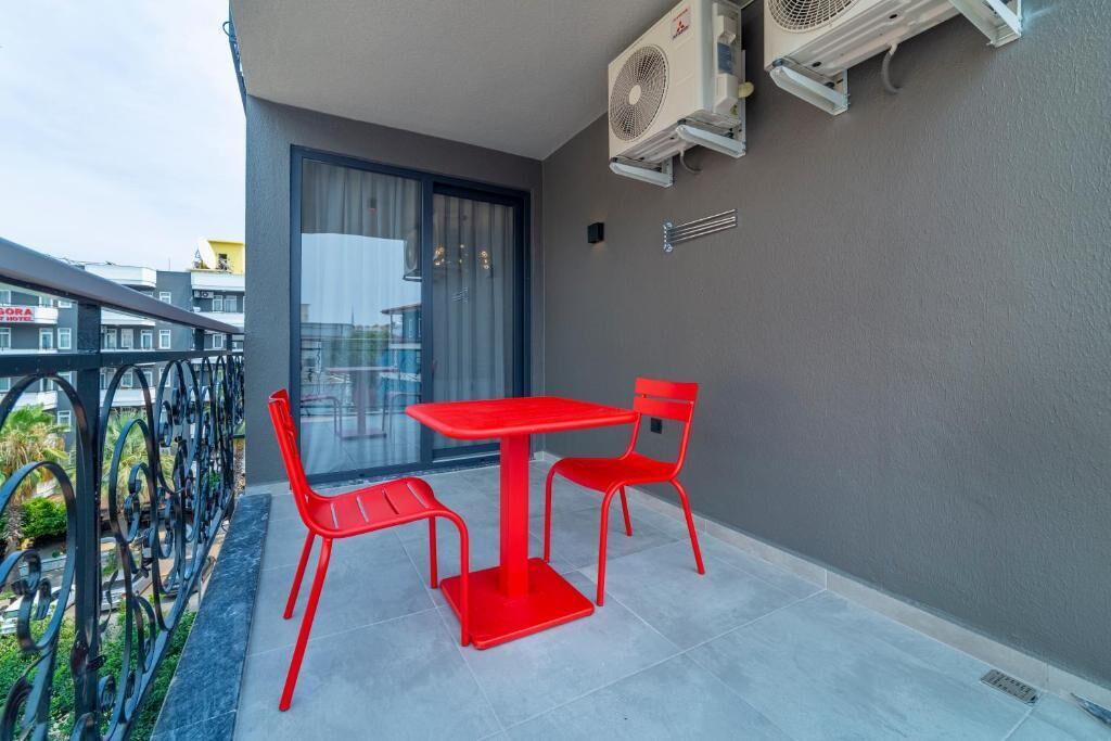A balcony with a red table and chairs, air conditioning units, and a view of a hotel in the background.