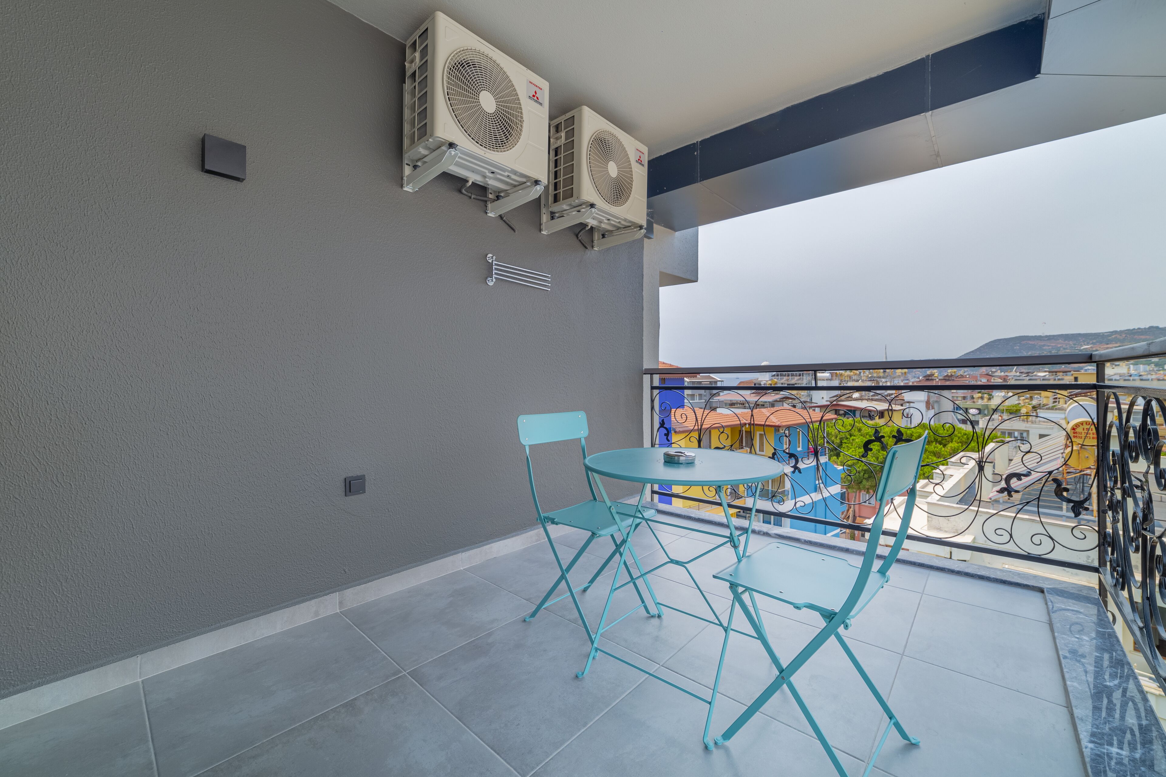 Un balcon doté de deux unités de climatisation, d’une petite table avec des chaises et d’où l’on peut admirer un bâtiment aux couleurs vives en arrière-plan.