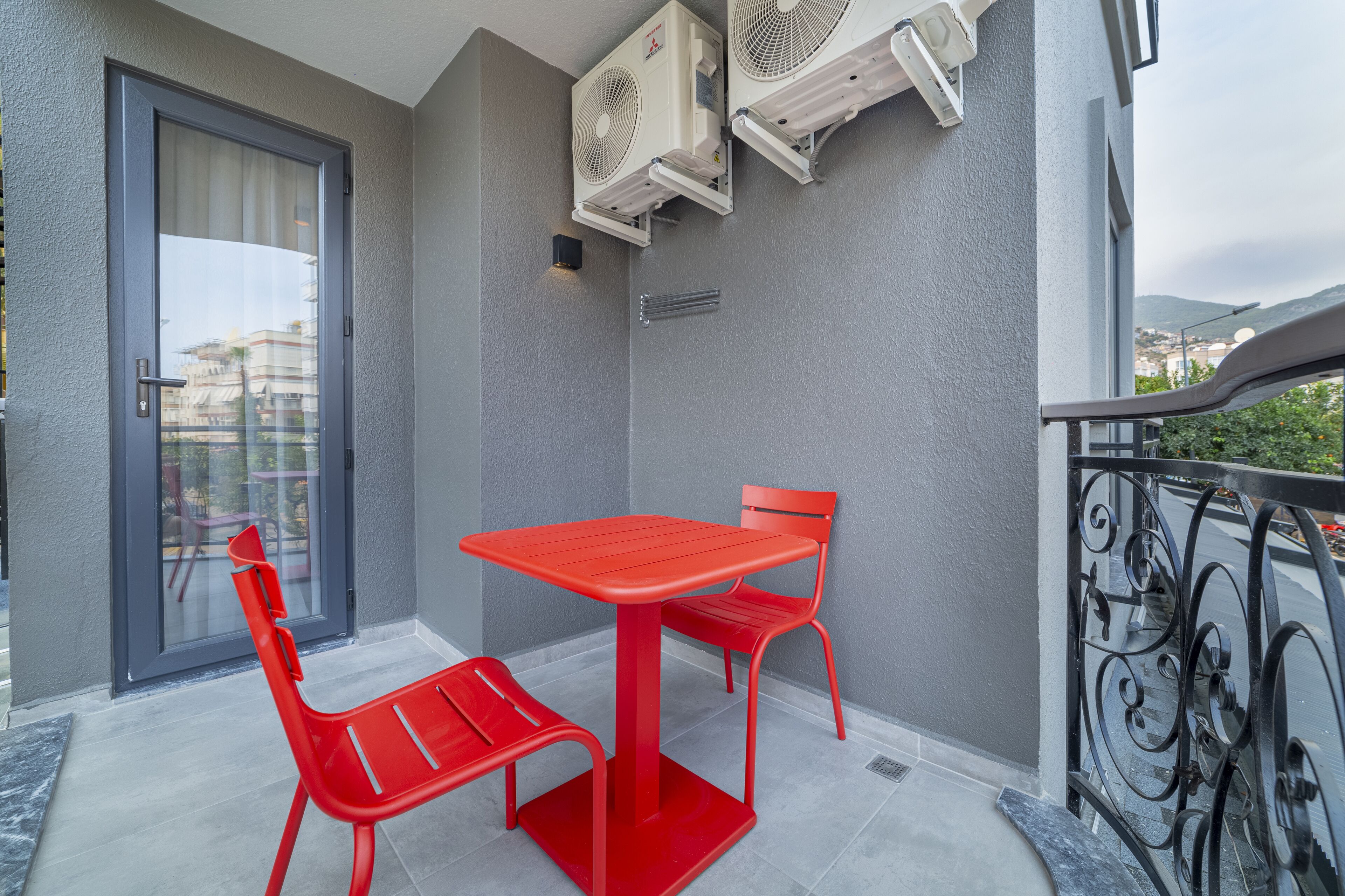 A balcony with a red table and chairs, air conditioning units, and a view of the city.