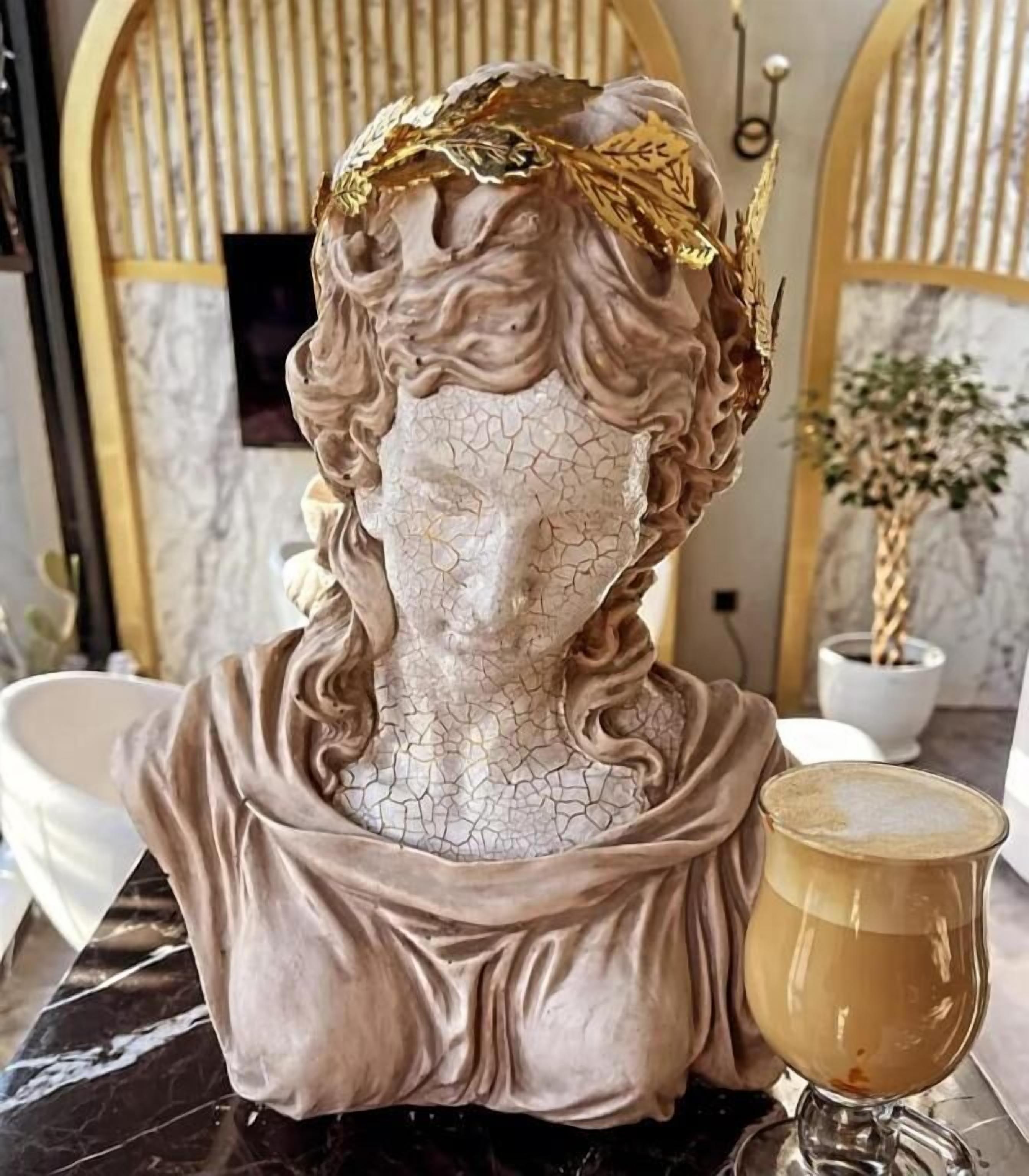 A bust sculpture with a golden leaf crown, placed on a marble surface next to a glass of coffee.