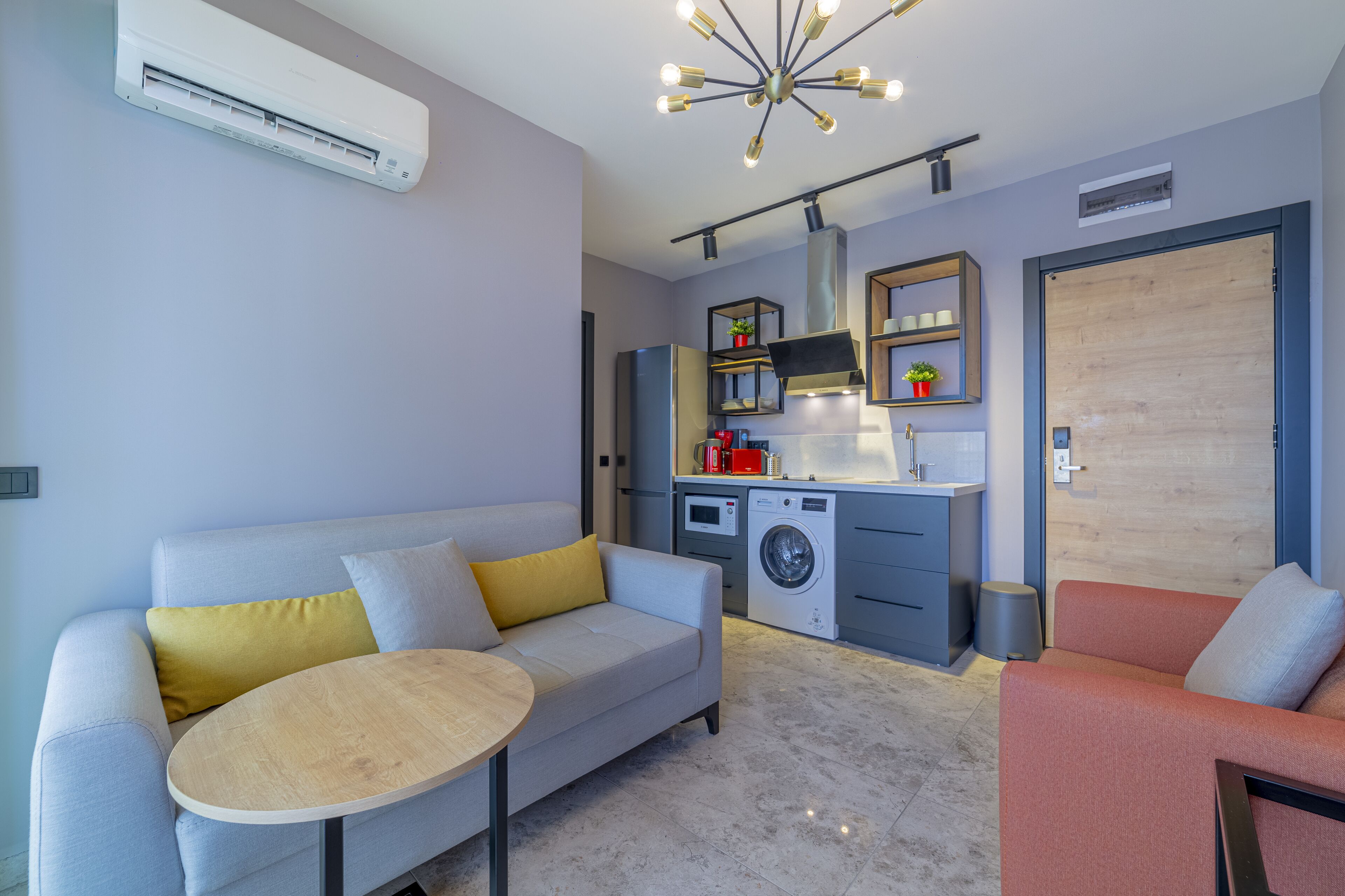 A modern apartment with a grey sofa, a round wooden table, a compact kitchen with a washing machine, and a wooden door.