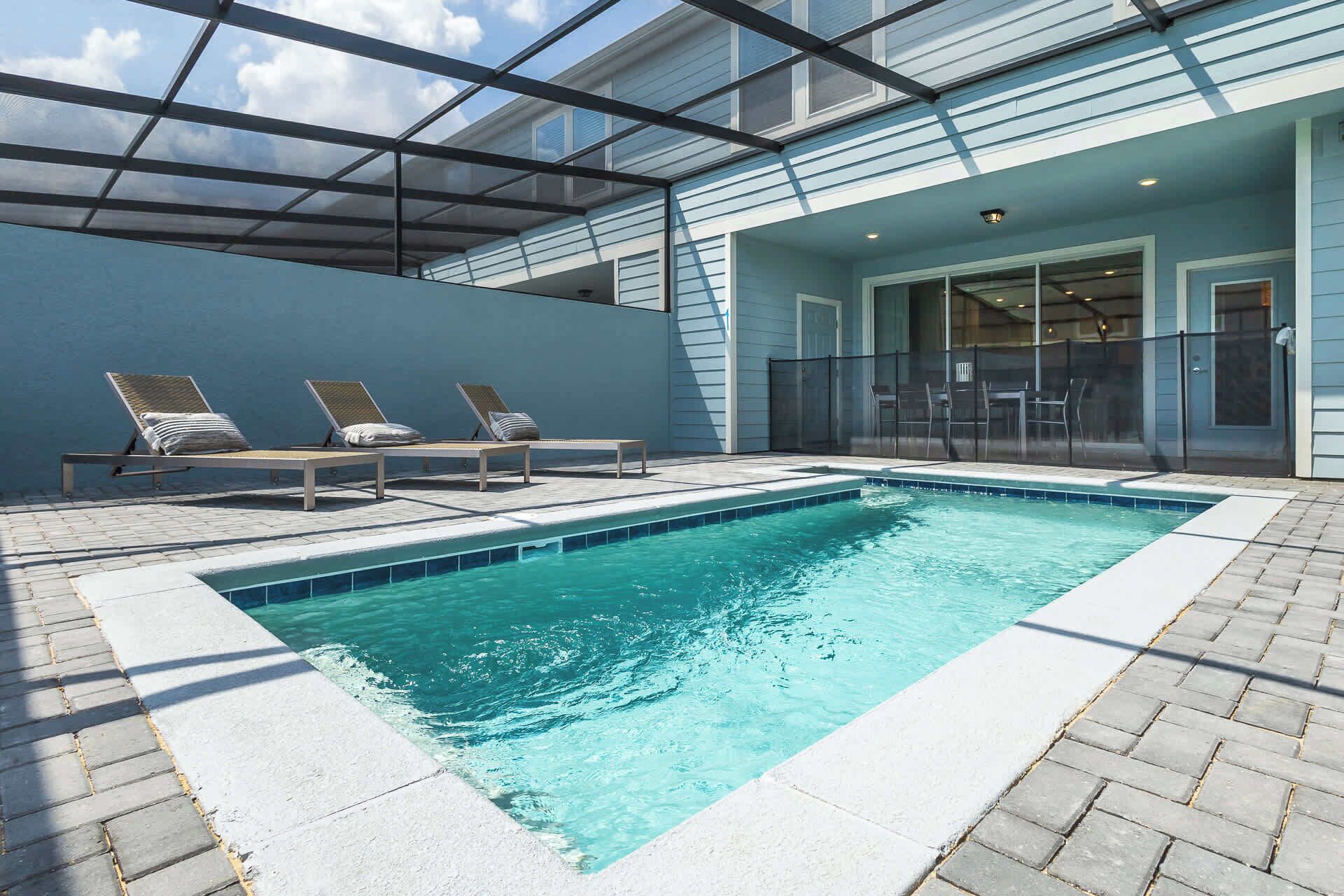A rectangular pool with a clear blue water surface, surrounded by a paved deck. There are three lounge chairs with cushions, and a glass-enclosed patio area with a table and chairs.
