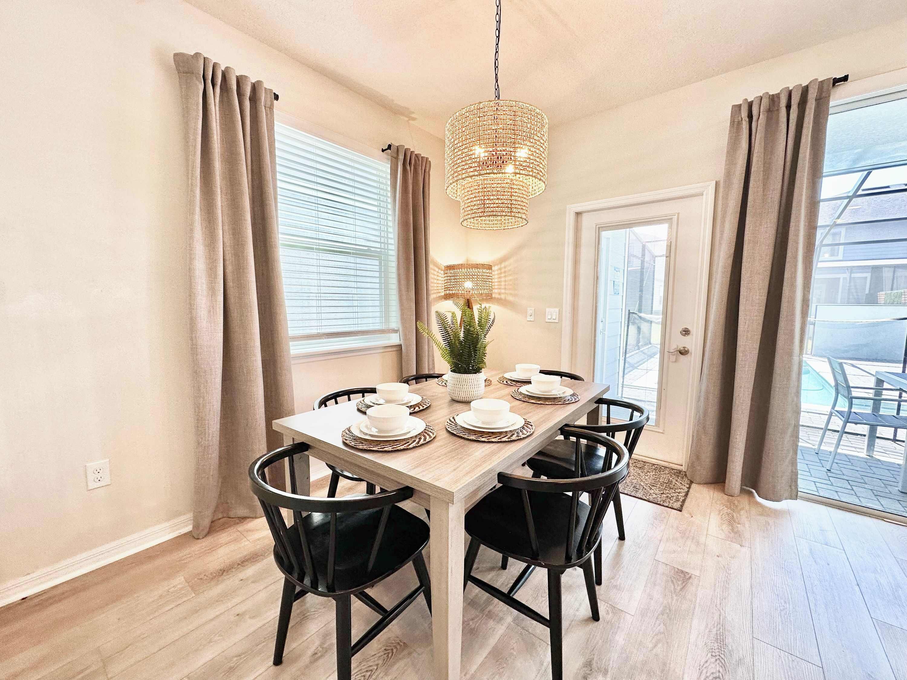 A dining area with a wooden table set for four, black chairs, a hanging chandelier, and a potted plant.