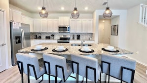 A modern kitchen with white cabinets, a stainless steel refrigerator, a microwave, and a double oven. There are four bar stools around a granite island with a sink and a faucet. - Adventure of a Lifetime! 5 BDR @ Windsor Island (Davenport)