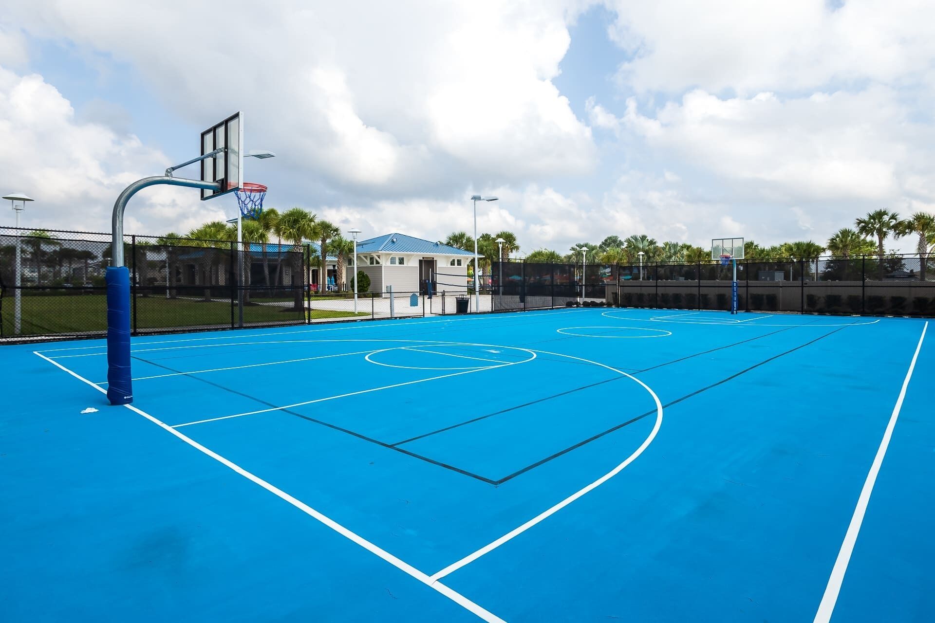 An outdoor basketball court with a blue surface, a basketball hoop, and a fence surrounding the area.